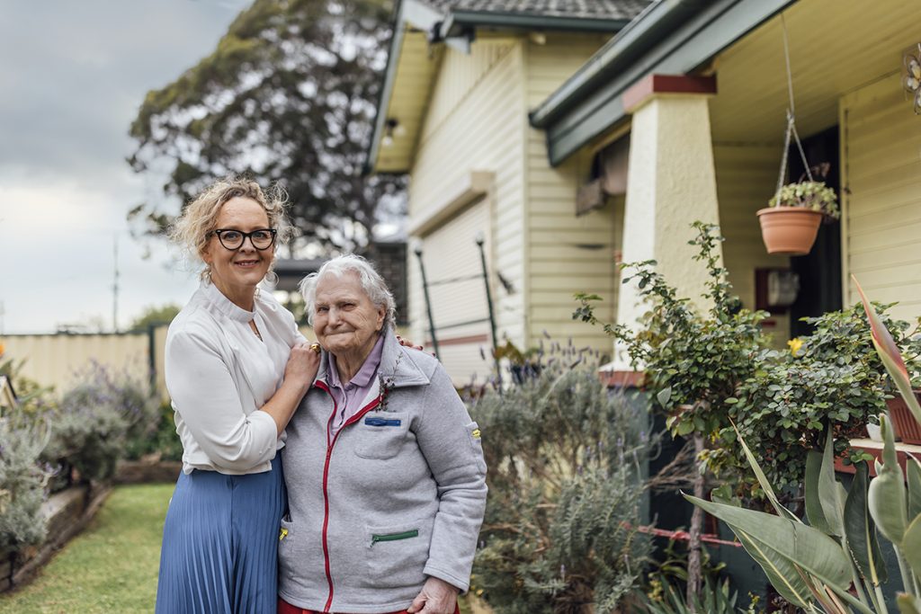 Two women outside old style home in front garden. One woman is elderly and the other is younger and standing next to her