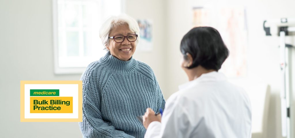 A senior woman sits up on the exam table at a doctors appointment. She is dressed casually in a blue sweater and has a smile on her face as she glances at the doctor. Her female doctor is wearing a white lab coat and seated in front of her as she takes notes on a tablet with Bulk Billing Logo
