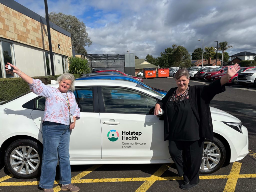 Two women smiling with arms up to camera stood beside new Holstep Health hybrid vehicle