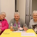 Three older women seated at yellow table