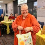 An older woman dressed in orange holds a gift bag and smiles at camera