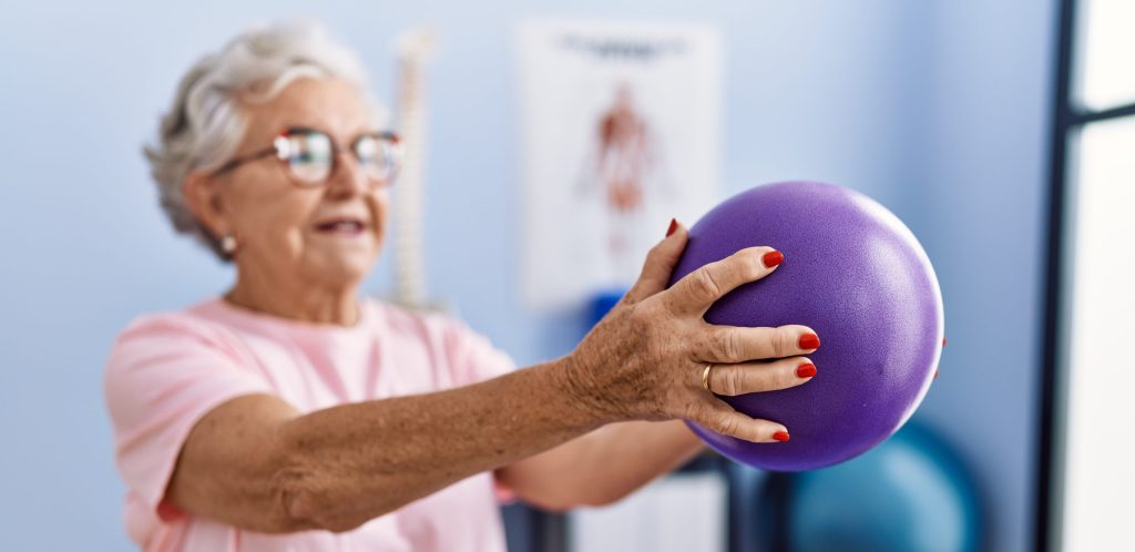 Senior grey-haired woman having rehab session using ball at physiotherapy clinic