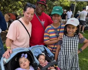 Two adults and two children smile at the camera beside a stroller carrying two toddlers with Down syndrome.