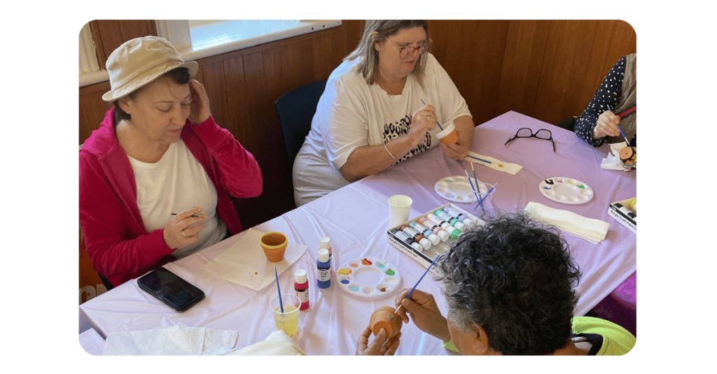 Three people sit round arts and craft table painting mini pots.